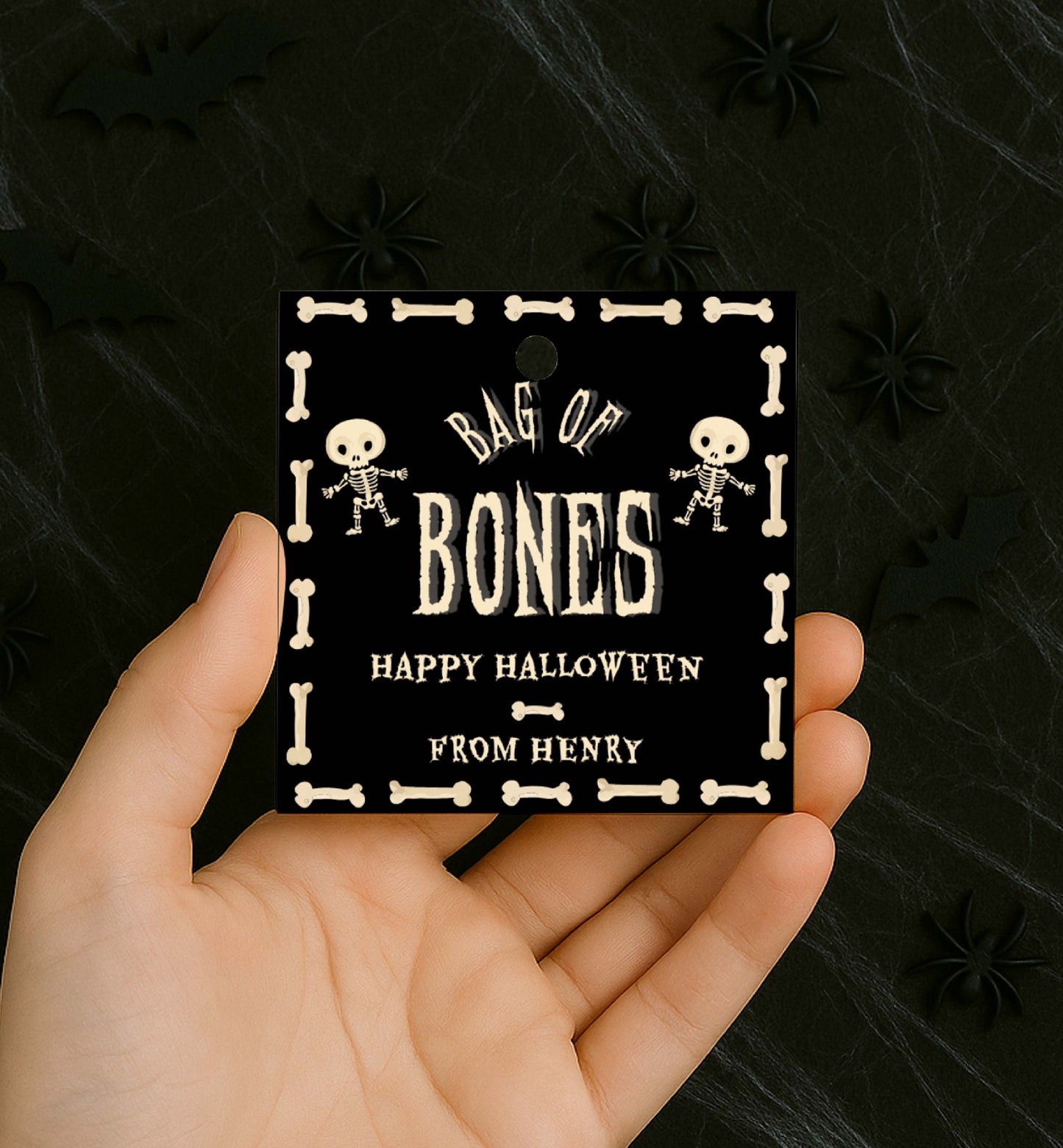 Hand holding a 'Bag of Bones' Halloween tag with skeleton designs on a dark background. Bag Of Bones Treat Tag, Skeleton Bones, Printable Halloween Favor Tags, Trick Or Treat Party Favors, Classroom Halloween Gift
