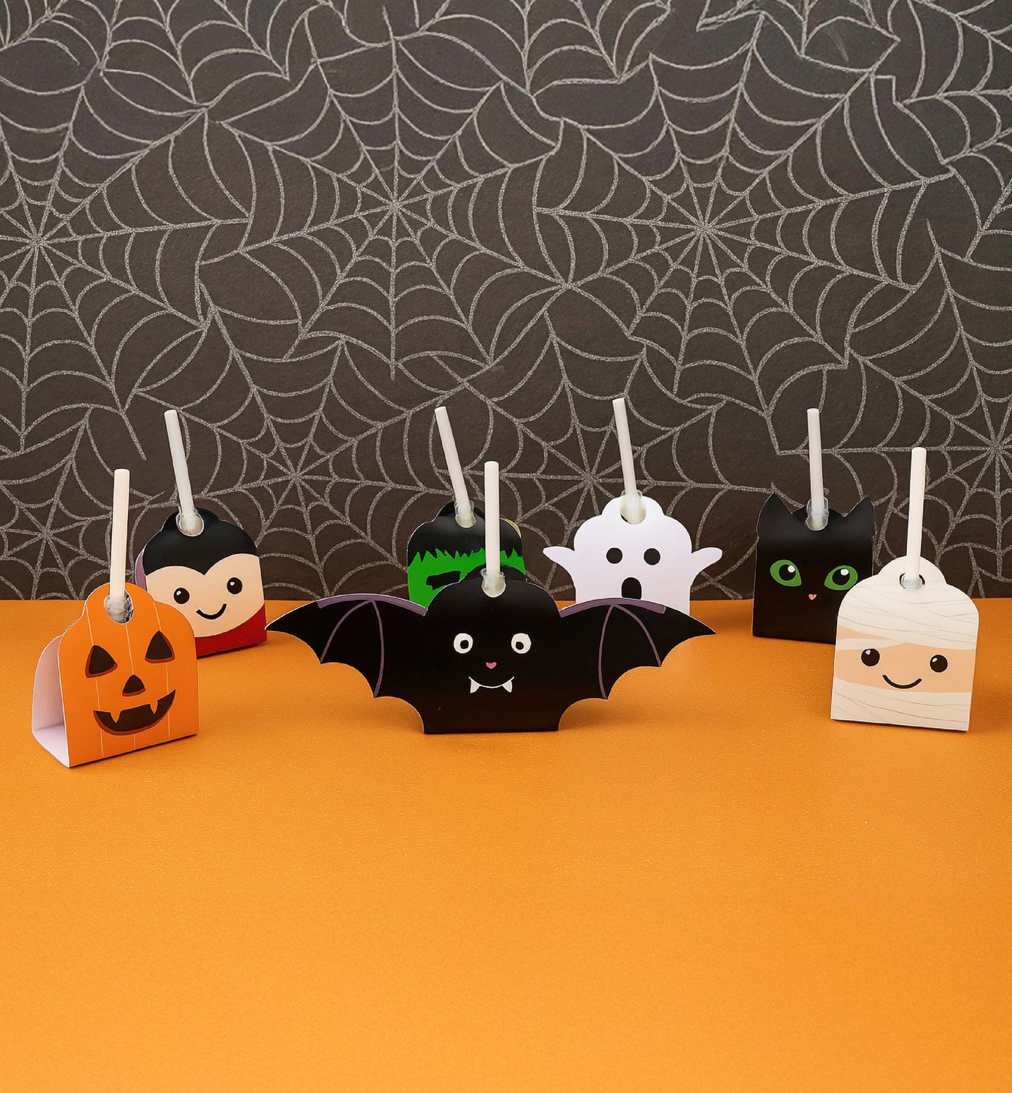 Halloween-themed paper decorations including a bat, pumpkin, ghost, and witch on an orange surface with spider web background. Printable Halloween Lollipop Tags Template, Halloween Trick or Treat Sucker Tag, Kids Classroom Halloween, Friends Halloween Gift