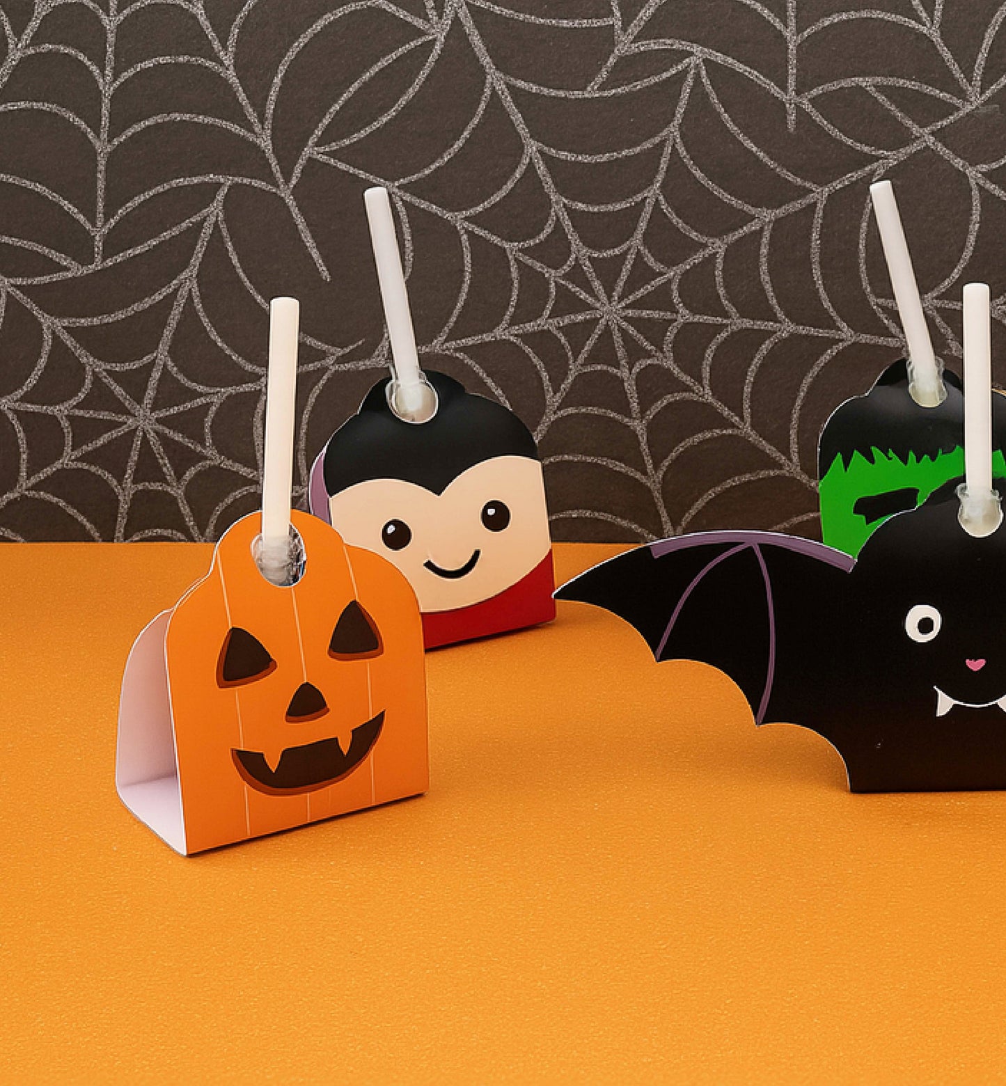 Halloween-themed paper decorations including a pumpkin, vampire, and bat on an orange surface with a spider web background. Printable Halloween Lollipop Tags Template, Halloween Trick or Treat Sucker Tag, Kids Classroom Halloween, Friends Halloween Gift