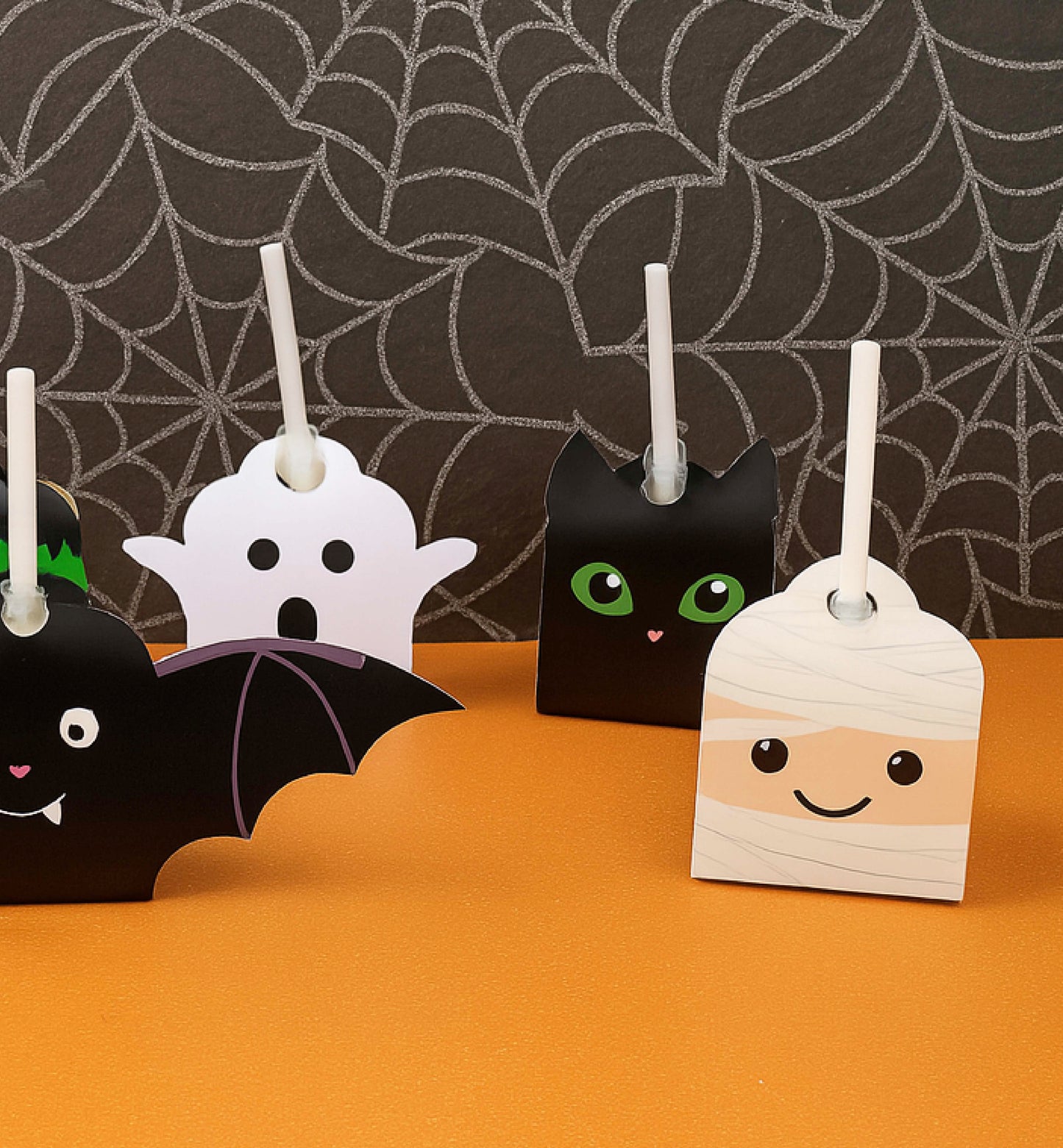 Halloween-themed candle holders shaped like a bat, ghost, and mummy on an orange surface with a spiderweb background. Printable Halloween Lollipop Tags Template, Halloween Trick or Treat Sucker Tag, Kids Classroom Halloween, Friends Halloween Gift