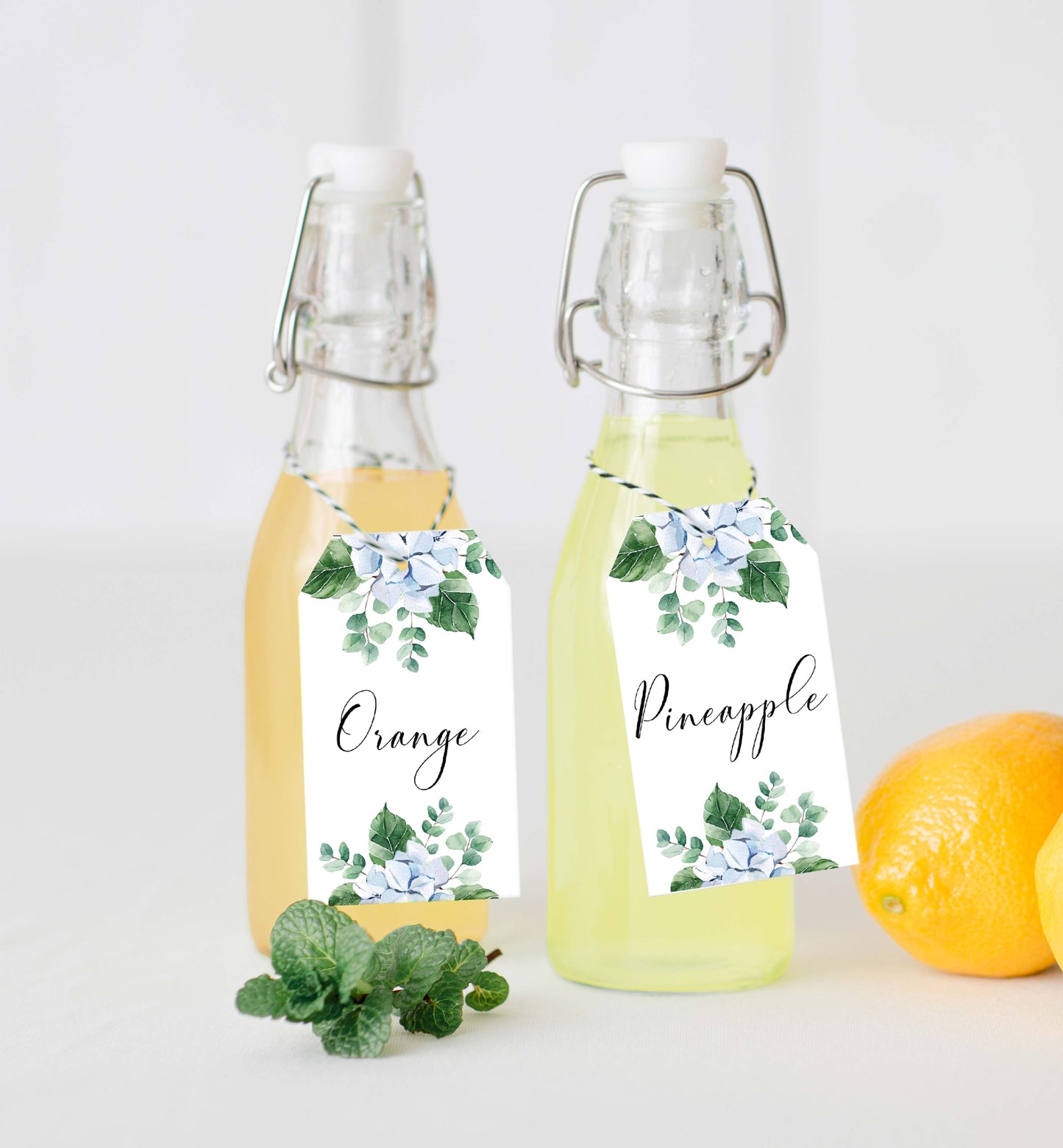 Two glass bottles with floral labels, one labeled 'Orange' and the other 'Pineapple', on a white background. Printable Mimosa Bar Sign and Juice Tags Template, Blue Hydrangea Boy Baby Shower Drinks Bar Menu Sign, Gender Neutral, Ferras Blossom