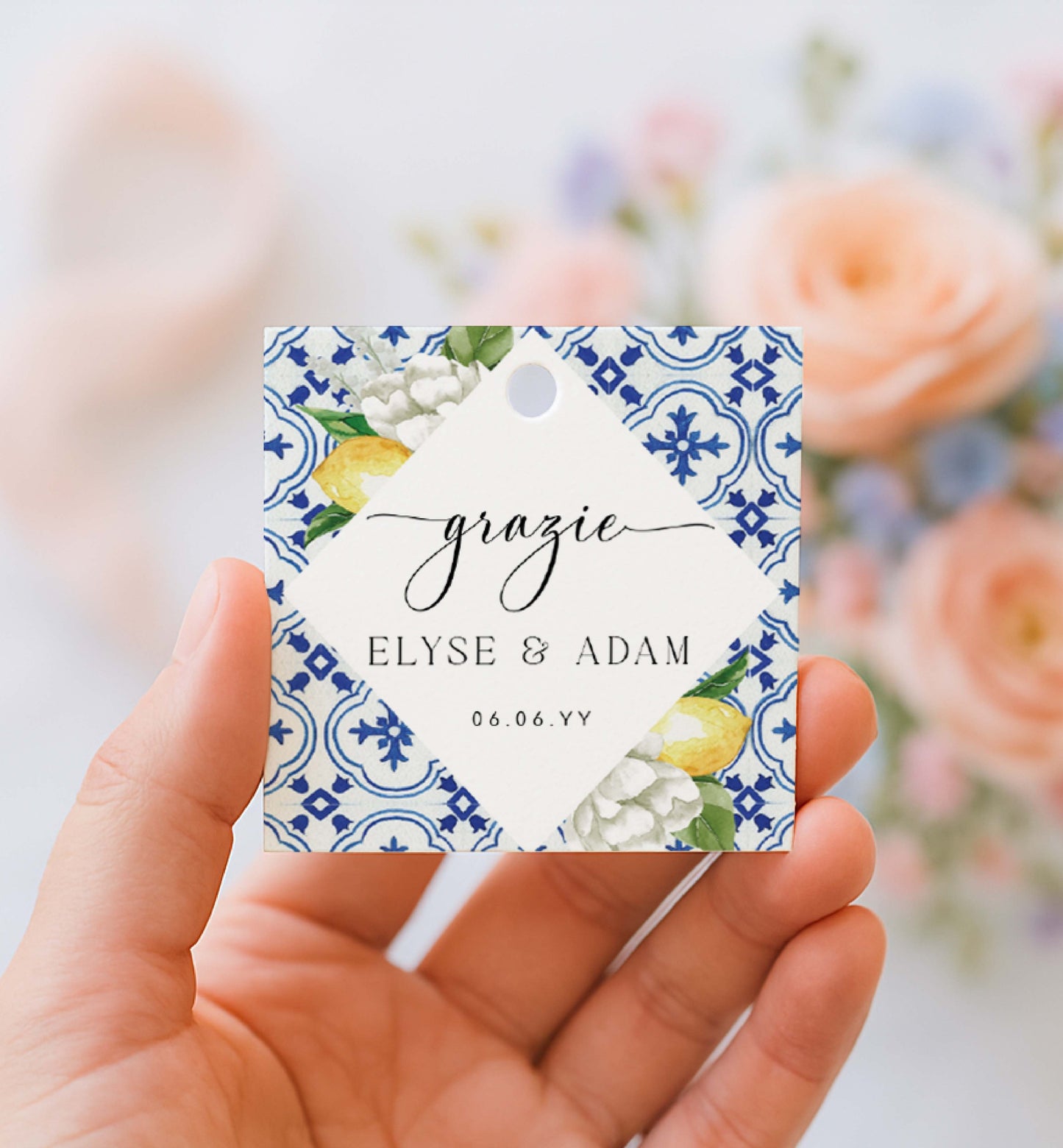 Hand holding a decorative tag with floral design and text against a blurred floral background. Printable Thank You Tag, Mediterranean Blue Tile Favor Tags, Lemons Thank You Grazie Tags, Ευχαριστώ Favor Tags, The Med