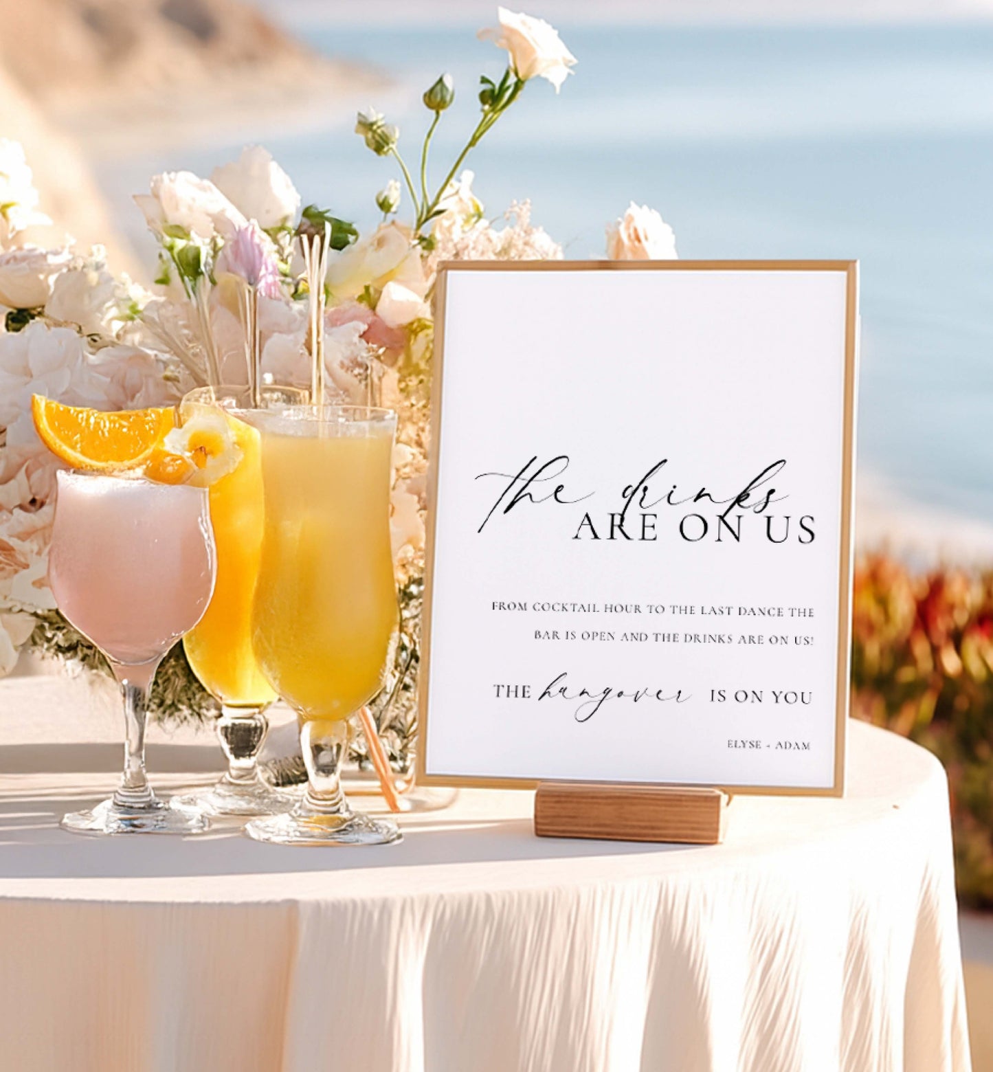 Table with cocktails and a sign reading 'The drinks are on us' against a scenic background. Printable Open Bar Sign Template, Minimalist Wedding Open Bar Sign, Printable Drinks Are On Us Sign, Wedding Bar Menu Sign, Ellesmere