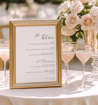 Gold-framed menu on a table with pink cocktails and flowers