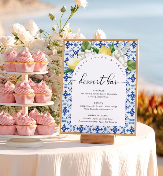Dessert table with pink cupcakes and a decorative dessert bar sign against a beach backdrop.