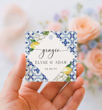 Hand holding a decorative tag with floral design and text against a blurred floral background. Printable Thank You Tag, Mediterranean Blue Tile Favor Tags, Lemons Thank You Grazie Tags, Ευχαριστώ Favor Tags, The Med