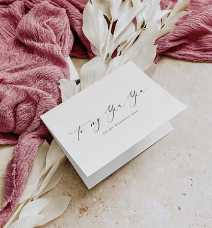 White card with cursive text on a textured surface with pink fabric and white flowers.